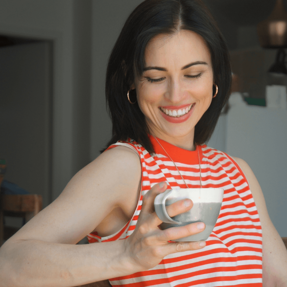 Farbfoto einer lächelnden Frau (Stillberatung Julia Lüders) mit dunklem Haar, die entspannt in einem rot-weiß gestreiften Oberteil sitzt und eine grau-weiße Kaffeetasse in der Hand hält.