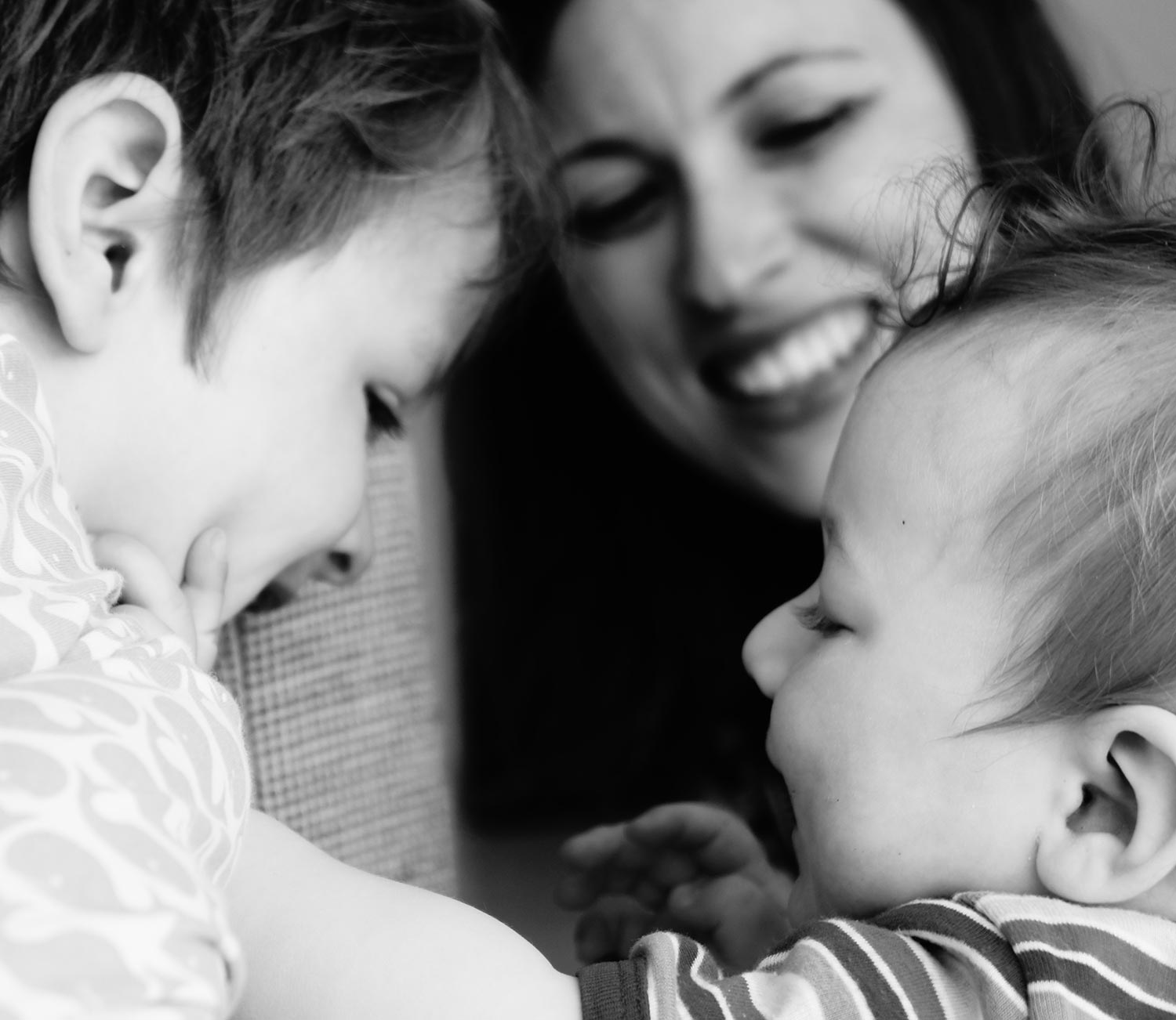Schwarz-weiß Nahaufnahme einer lächelnden Frau, die liebevoll auf ein Baby und ein Kleinkind blickt.