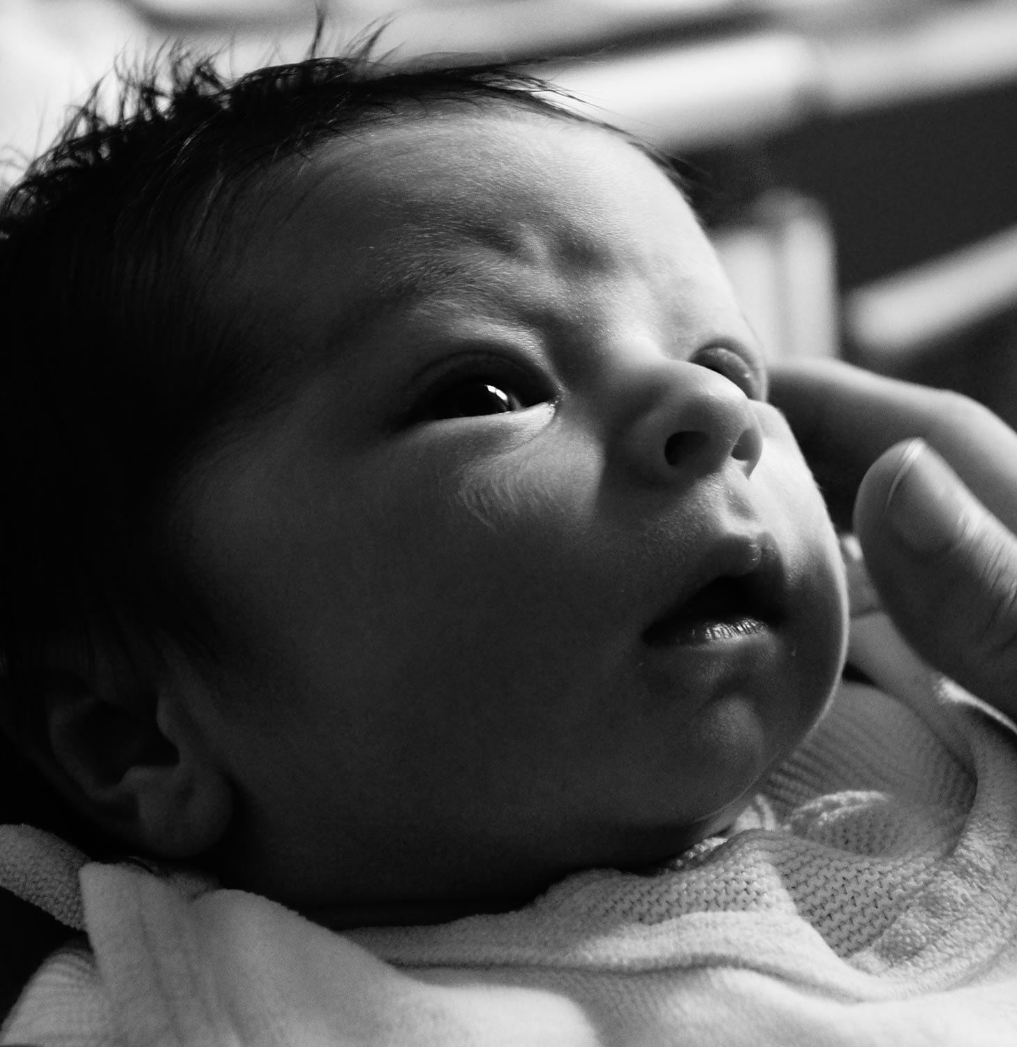 Schwarz-weiß Nahaufnahme eines neugeborenen Babys, das staunend nach oben blickt, während eine Hand sanft seinen Kopf berührt.